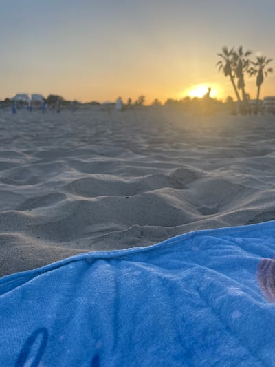 The image captures a low-angle view of a beach at sunset. In the foreground, a textured blue towel lies on rippled sand. The background is blurred, showing a hazy horizon with silhouetted palm trees and a few indistinct figures on the beach. The sun is setting, casting a warm, golden glow across the sky. The location appears to be a public beach, likely in Canet d'en Berenguer, Spain, given the presence of palm trees and the typical ambiance of a Mediterranean sunset. The scene is serene, focusing on the natural beauty of the beach and the approaching evening.