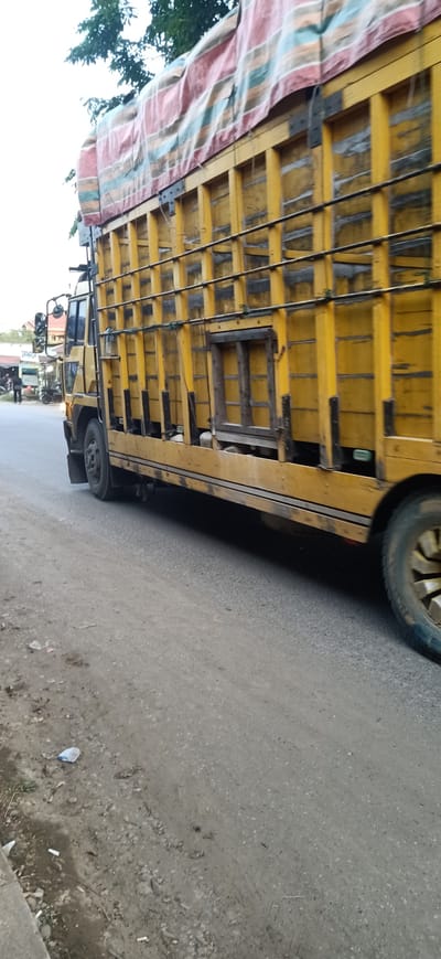 This image shows a yellow truck with a covered bed driving down a road in Gampongteungoh, Indonesia. The truck's bed is covered with a tarp that has horizontal stripes of red, white, and green. The truck is in motion, as indicated by the slight blur of the wheels. The scene appears to be a rural or semi-urban area, with some buildings and trees visible in the background. The road surface is paved and appears to be in good condition. There is also some dirt and debris on the side of the road. The lighting suggests it is daytime, possibly late morning or early afternoon. There are no people prominently featured in the image, though some figures might be discerned in the distance on the left side of the frame. The overall impression is of a typical day in a Southeast Asian setting, with the truck likely involved in transportation or logistics. No visible text is present in the image.