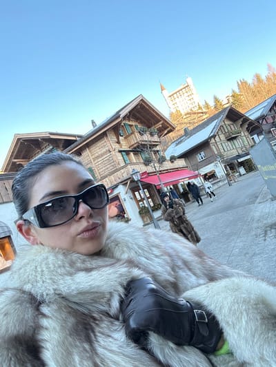 A woman is depicted in a close-up, chest-level selfie outdoors on a clear day in Gstaad, Switzerland. She faces the camera, wearing large black sunglasses and a light-colored fur coat, with her left hand, gloved in dark leather, resting on her chest. Her lips are slightly pursed. In the mid-ground, a paved pedestrian street is visible, flanked by traditional Swiss chalet-style buildings with wooden facades, balconies, and gabled roofs, some lightly dusted with snow. One building features a red awning over a storefront. Several other individuals are present in the mid-ground, including one person in a brown fur coat holding a phone. In the distant background, atop a wooded hill, the prominent white structure of the Gstaad Palace hotel with its distinctive pointed tower is visible under a clear blue sky. The overall scene suggests a winter or late autumn setting.