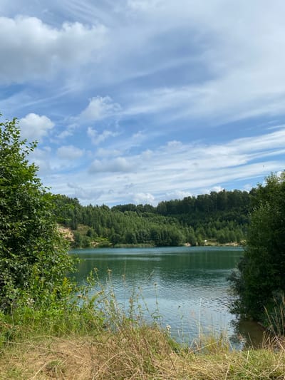 The image depicts a serene natural landscape at the Plintovka Lake in Plintovka, Russia. The foreground is dominated by dry grass and a sandy bank, leading the eye towards the shimmering turquoise water of the lake. The lake itself is calm, reflecting the blue sky above. Across the water, a dense, green forest covers rolling hills. The sky is a vibrant blue, dotted with scattered white clouds, some of which appear wispy and streaky, suggesting a light breeze. On the far shore, a few small figures are visible, possibly picnickers or swimmers, along with a couple of white tents, indicating a recreational area. The overall impression is one of a peaceful, sunny day in a picturesque natural setting.