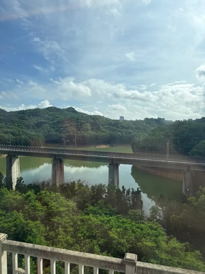 The image depicts a wide shot of a body of water with a bridge spanning across it. The water is calm and reflects the sky and surrounding greenery. The bridge is a substantial structure supported by concrete pillars and carries railway tracks. Lush green hills covered in trees rise behind the water, with a few tall buildings visible in the distance on the crest of one of the hills. The sky above is partly cloudy, with patches of blue visible between fluffy white clouds. In the foreground, a white railing suggests the viewpoint is from an elevated structure, possibly a balcony or another bridge. The scene conveys a sense of natural beauty and infrastructure coexisting, suggesting a location that balances urban development with the environment. The location is identified as Longhua District, China.
