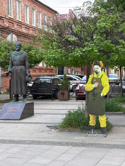 In Akhieli, Georgia, a bronze statue of a man in a long coat stands near a tree and a painted figure of a man in a yellow outfit and dark apron holding an object. A brick building and parked cars are in the background. The area is paved with light gray stones.