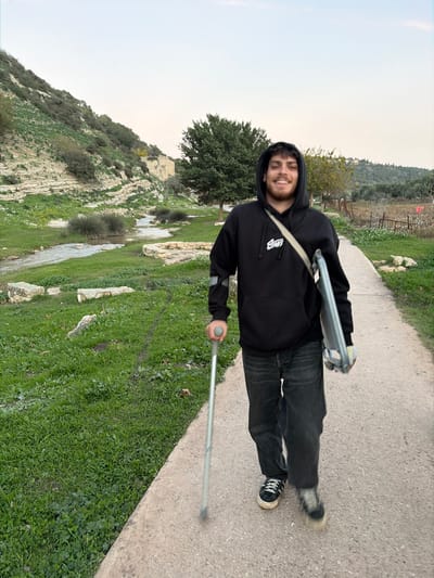 A young male, appearing to be in his late teens or early twenties, walks forward on a light-colored paved path. He has short dark hair, light facial hair, and is smiling with his mouth open. He wears a black hooded sweatshirt featuring a white logo on the chest, dark wash jeans, and dark sneakers with white laces. He holds a silver forearm crutch in his right hand. A silver-grey metallic object, possibly a scooter frame, is slung over his left shoulder with a beige strap.
The scene is an outdoor environment within the Emek Izrael Regional Council, Israel. To the left of the paved path, a grassy area with green vegetation gives way to a shallow, winding stream. Beyond the stream, a steep, rocky hillside rises, exhibiting distinct horizontal geological strata and covered with sparse green shrubbery. A light-colored stone building or ruin is visible on this hillside in the mid-background. Along the stream's edge, partial stone structures or retaining walls are present. To the right of the path, an agricultural area with rows of small trees, consistent with an olive grove, extends towards a distant, tree-covered hill. A large, full-leafed tree stands centrally in the mid-ground. The sky above is a uniform, light grey-blue.