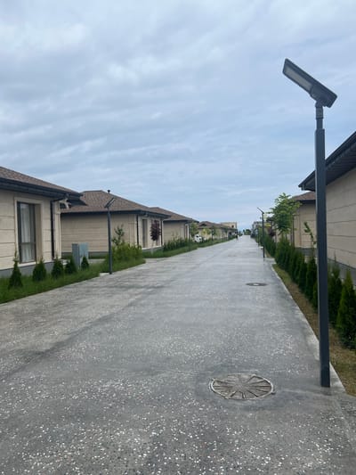The image depicts a street in the city of Bosteri, Kyrgyzstan. The street is paved with concrete and lined with identical, modern houses on both sides. The houses have beige exteriors with dark tiled roofs and visible windows. There are small, manicured cypress trees planted along the front of the houses. Streetlights are positioned at intervals along the road. The sky is overcast with gray clouds. The scene suggests a residential area that is well-maintained and uniform in design. There are no visible people in the foreground or midground, though a distant figure can be seen walking further down the street.