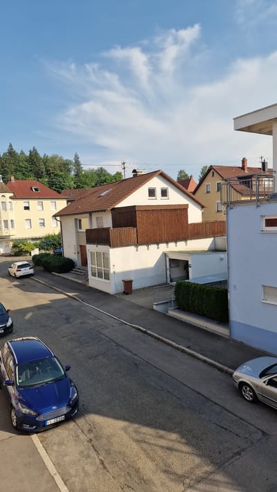 The image shows a street in Villingen-Schwenningen, Germany, on a sunny day with some clouds. On the left side of the street, there is a white Volkswagen Golf parked. Further down the street, a blue Ford Focus is parked. Across the street, there are several houses. The house in the foreground on the right is light blue, and has the number '8' painted on the side. Next to it, a white house with a brown wooden balcony is visible. Behind these houses, more houses with red-tiled roofs and green trees are visible. The sky is blue with scattered white clouds. The scene appears to be a residential area.