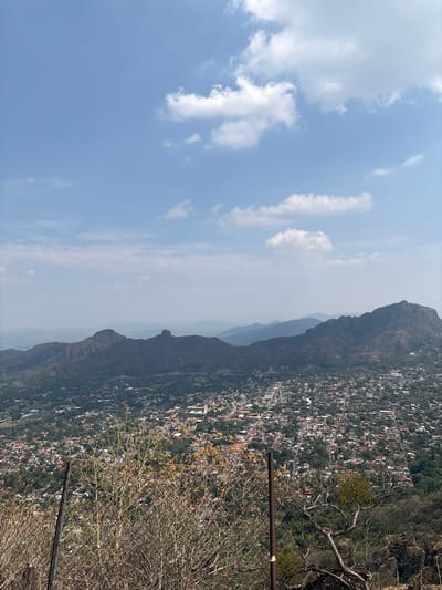 The image shows a view of Tepoztlán, Mexico, from an elevated vantage point, possibly a mountainside. The foreground features dry vegetation and a metal fence. The middle ground shows the sprawling cityscape nestled amidst hills and mountains, with the distinct Tepozteco mountain visible. The background includes a blue sky with scattered white clouds.