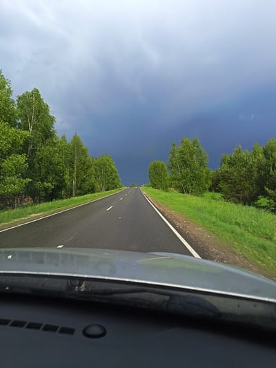 The image depicts a view from inside a vehicle, likely a car, driving on an asphalt road. The road is flanked by green trees and grass. The sky above is dark, possibly indicating an approaching storm. The location is Aktushevo, Russia. There are no people visible in the image. The scene is outdoors.