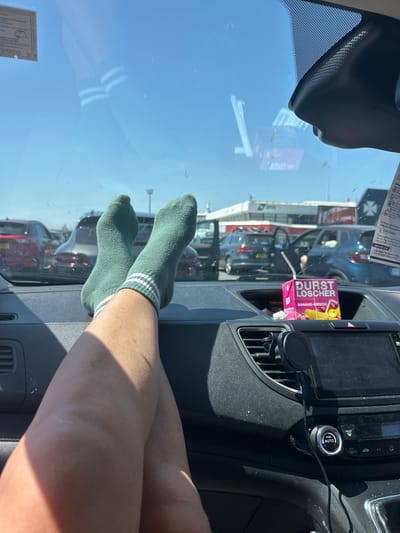 A view from inside a car shows a pair of legs resting on the dashboard and windshield. The legs are wearing green socks with white stripes. A juice box labeled "Durst Löscher Banane-Kirsch" is in the car's dashboard area, along with a straw. Outside the windshield, several parked cars and buildings are visible, suggesting a parking area or city street. Based on the presence of the "DFDSUKUpdates" and "www.dfds.com" texts, it is likely close to ferry terminal in Loon-Plage, France.