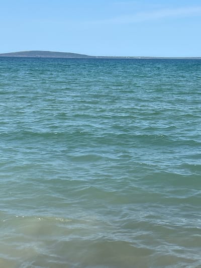 The image shows a wide expanse of calm, turquoise ocean water under a clear blue sky. In the distance, a low-lying island or peninsula with a dark, undulating silhouette is visible on the horizon, separating the water from the sky. The foreground of the image displays the gently lapping waves at the edge of the water, with the sandy seabed visible beneath the surface. The scene evokes a sense of tranquility and natural beauty. This location is consistent with the coastal landscape of Ballybunnion, Ireland, known for its scenic beaches and ocean views. There are no people or specific structures visible in the frame, emphasizing the natural environment.