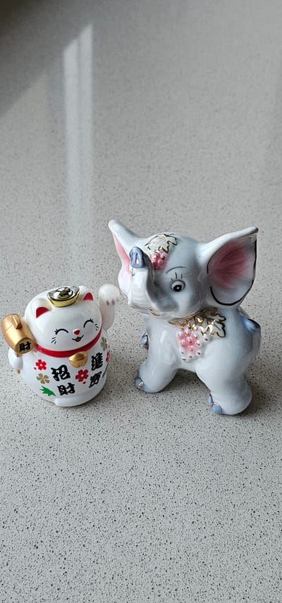A maneki-neko figurine and a light blue ceramic elephant figurine are placed side-by-side on a speckled grey countertop. The maneki-neko, on the left, is white with red ears and a red collar adorned with gold bells and floral patterns. It holds a gold mallet in its left paw and has Chinese characters on its body. The elephant, on the right, is a light blue color with pink inner ears, and gold accents on its trunk, ears, and a cluster of pink grapes on its chest. The background is a blurred grey surface, suggesting an indoor setting. The image composition is a medium shot, focusing on the two figurines. There is a bright rectangular light reflection on the countertop behind the figurines. There is no indication of the location being Borovoye Matyushino, Russia.
