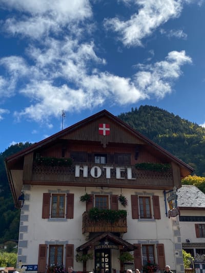 This image displays the Hotel Les Touristes, located in Abondance, France, under a bright blue sky with scattered white cumulus clouds. The building exhibits a traditional Alpine architectural style, featuring a prominent dark brown wooden gabled roof with overhangs. A red shield with a white Swiss cross is affixed to the wooden gable, and a television antenna protrudes from the roof peak.
The upper section of the hotel's facade is constructed of dark brown wood, featuring a balcony with intricately carved wooden railings and window boxes containing pink flowers. Large white dimensional letters spelling "HOTEL" are mounted on this balcony's railing. Below, the building transitions to light-colored (off-white) rendered walls, accentuated by grey stone quoins at the corners. Multiple windows with brown wooden shutters are visible across these lower floors.
The main entrance is positioned beneath a small, covered awning, above which a smaller balcony adorned with flowers is situated. A sign bearing the name "LES TOURISTES" is displayed above this entrance. Additional hanging flower baskets are suspended from balconies and window areas. A black metal lantern is affixed to the left corner of the building's facade. Partially visible at the bottom of the frame are blurred figures of individuals and portions of parking signage. In the background, steep, forested mountains rise, displaying a mix of green and some yellow foliage, indicative of late summer or early autumn.
