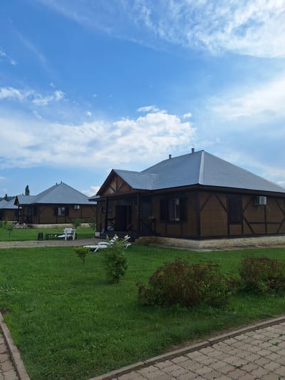 In Atamanovka, Russia, a resort complex features several identical, rustic wooden cabins with dark metal roofs. The cabins are arranged in a row, with the nearest one to the right featuring a porch with dark wooden railings and a dark door. The cabins have wooden siding with dark timber framing accents and stone bases. Each cabin is equipped with an outdoor air conditioning unit.
The grounds are covered in lush green grass, with a paved walkway made of light grey interlocking stones bordering the area. Several white lounge chairs are scattered on the lawn in front of the cabins, some with umbrellas. Small, manicured bushes and trees are strategically placed throughout the landscape. In the distance, more cabins are visible, as well as a treeline and hills under a bright blue sky with scattered white clouds. The atmosphere appears calm and serene, suggesting a leisurely retreat.
