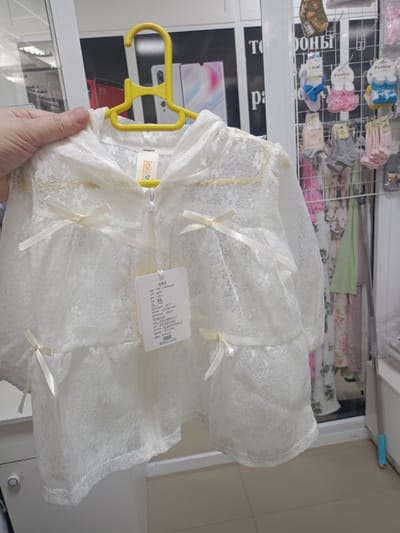 A hand is holding a white lace jacket with bows. The jacket is hanging on a yellow hanger, and there is a tag attached to it. The background shows a rack of clothes, including socks and other garments. The setting appears to be a retail store, possibly a clothing boutique, given the display of merchandise. Some text is visible on the tag, including "Jadventure," "XL," and various Chinese characters. There is also text on the background display which appears to be "Pamily" and other Russian lettering. The lighting suggests an indoor environment.
