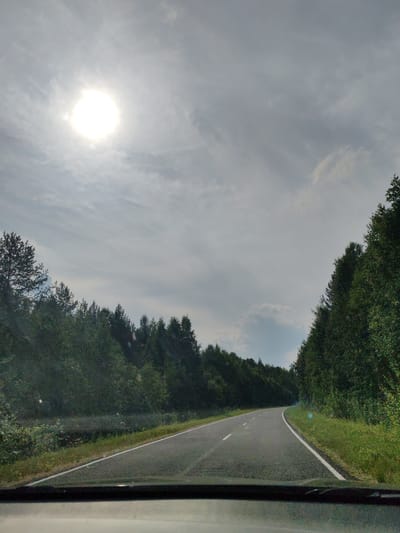 The image depicts a road winding through a forest under a cloudy sky with the sun shining brightly through a gap in the clouds. The road appears to be paved and has white lane markings. Dense green trees line both sides of the road, forming a canopy that partially obscures the sky. The sun, a bright white orb, is positioned in the upper left quadrant of the image. The foreground shows the interior of a vehicle, suggesting the image was taken from a car. The scene is consistent with a rural or forested area. The provided context states the location is the city of Chukhlem, Russia, which is a small town surrounded by natural landscapes.