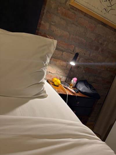 The image captures an intimate interior scene, likely a bedroom, focusing on a bed, a brick wall, and a bedside arrangement. A large bed with white sheets and pillows dominates the foreground and right side of the frame, creating a sense of comfort and coziness.
To the left, an exposed, reddish-brown brick wall provides a rustic backdrop. On this wall, a framed artwork is visible in the upper left quadrant. It features a simple black line drawing, possibly of a human figure, and bears a signature that appears to be "müller" or similar. Below the artwork, a black, articulated desk lamp is positioned, emitting a bright, focused beam of light that creates a starburst effect as it shines towards the wall and a wooden shelf below it.
On this wooden bedside shelf, several items are arranged: a black bag, a bottle with a pink cap (possibly a cosmetic product), a hairbrush with a pale handle, and a small cluster of bright yellow lemons. Wires are also visible on the shelf, suggesting the presence of electronic devices. A dark, possibly black, piece of furniture or a headboard is partially visible behind the shelf and lamp.
The lighting in the room is dim overall, suggesting it is nighttime or a low-light setting, with the lamp providing the primary illumination and contributing to a warm, inviting atmosphere. The brick wall contrasts with the clean white bedding, implying a blend of rustic and modern aesthetics. No specific activity is taking place. The scene is set within the Overstrand Local Municipality, South Africa.