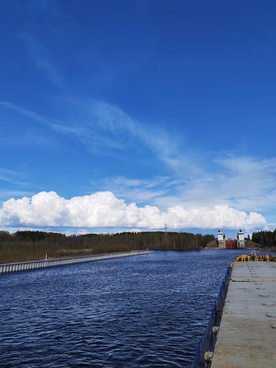 The image depicts the Volga–Baltic Waterway near Анхимово, Russia. A body of water fills most of the foreground, its surface rippled with gentle waves. A long, grey concrete structure extends from the left towards the center, appearing as a narrow embankment or pathway. Beyond the water, a line of trees forms a dense forest along the horizon. A power transmission tower is visible in the middle distance. In the background, a lock structure with towers is visible. The sky occupies the upper half of the frame, displaying a bright blue hue with wispy cirrus clouds at the top and a band of puffy cumulus clouds along the middle. The image is taken from an elevated position, likely from a vessel, and appears to be captured during daylight under clear weather conditions.