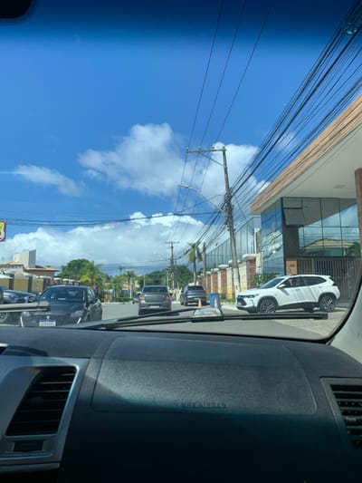 The image depicts a street view captured from within a stationary vehicle, looking northeast on R. Gerino de Sampaio, at the intersection with R. Gerino de Sampaio, 255, in Pitangueiras, Lauro de Freitas, Bahia, Brazil. The foreground features the dark dashboard, air vents, and windshield wipers of the camera vehicle. The sky above is clear and blue with scattered white cumulus clouds. Numerous overhead utility poles are densely strung with a complex network of power and communication lines that cross the frame.
On the paved asphalt road, multiple vehicles are present. A dark gray Peugeot 208, bearing Brazilian Mercosul license plate SIK6G04, is positioned in the immediate left lane, facing the camera. Further down the street, a gray sedan and a dark blue SUV are visible, facing away from the camera in what appears to be a traffic lane. To the right, a white Chevrolet Tracker SUV is situated near the entrance of the "Nouvelle Lauro de Freitas Clínica e Centro Odontológico," a modern building with large glass windows and light-colored fascias. A single occupant is discernible in the driver's seat of the white SUV. A small, orange traffic cone or marker is visible on the road surface directly ahead of the camera vehicle. The broader environment includes additional low-rise buildings and abundant green foliage, including palm trees, bordering the street. No pedestrians or other identifiable individuals are visible in the scene.
