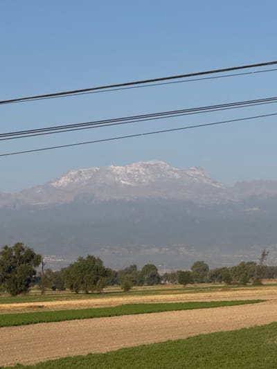 This image depicts an expansive outdoor scene in Santa Ana Xalmimilulco, Mexico, under a clear, light blue sky. Three parallel, dark-colored power lines traverse horizontally across the upper third of the frame. In the distant background, a prominent mountain range, consistent with Iztaccihuatl, spans the horizon; its upper elevations are distinctly covered in white snow, while its lower slopes appear veiled by a gray-blue atmospheric haze. The midground features a discontinuous line of scattered trees with varying green foliage. Behind these trees, faint, dispersed structures indicative of a distant settlement are subtly visible. The foreground is composed of agricultural fields divided into distinct horizontal sections. These sections vary: some consist of light brown, bare earth; others display vibrant green, low-lying crops; and a few show dry, yellow-brown vegetation. No individuals, animals, or direct human actions are observable within the frame. The environment is rural, showcasing agricultural land and a significant natural landmark.
