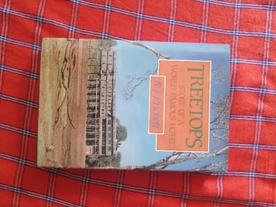 The image shows the cover of a paperback book titled "Treetops: Story of a World Famous Hotel" by R. J. Prickett. The book cover depicts a photograph of a large, wooden structure built high above the ground, seemingly in a forested area. The structure is made of a series of interconnected platforms and walkways, resembling a treehouse. There are trees surrounding the structure, and the ground beneath appears to be a grassy area with a few fallen branches. The book is lying on a red and blue plaid fabric, likely a blanket or tablecloth. The image does not contain any information about the exact location or any people.