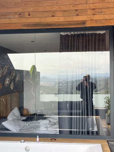 This is a vertical, interior shot of a modern room with a large glass wall, likely a hotel room or a luxurious home. Through the glass, a landscape is visible, featuring rolling green hills with some scattered buildings, suggesting a rural or semi-rural location. The sky is bright and appears to be daytime, possibly late morning or early afternoon given the lighting.

Reflected in the glass are two individuals. In the upper portion of the reflection, a person wearing dark clothing and a cap is holding a camera and appears to be taking a photograph of the landscape. Their posture suggests they are focused on their activity. In the lower portion of the reflection, another person, possibly female, is lying on a bed. They are wearing a bikini or swimwear.

Inside the room, visible through the glass, is a bed with rumpled white sheets and pillows, indicating recent use or a relaxed atmosphere. There are also decorative elements on the wall, including woven baskets and other patterned wall hangings, adding a bohemian or artisanal touch to the decor. The room appears to be well-lit, with natural light streaming in. The overall mood is one of leisure and relaxation, possibly a vacation setting. The wooden accents on the exterior wall contrast with the modern glass and interior design.