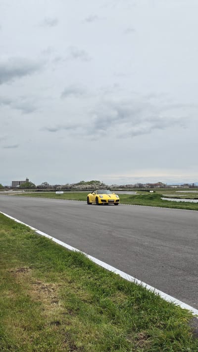 The image shows a yellow car on a racetrack at the Serres Racing Circuit, located near the village of Kaloyanovo, Bulgaria. The car is positioned on the asphalt track, with a white line marking the edge. Green grass borders the track on the right, while the left side presents a view of trees, buildings, and a cloudy sky.