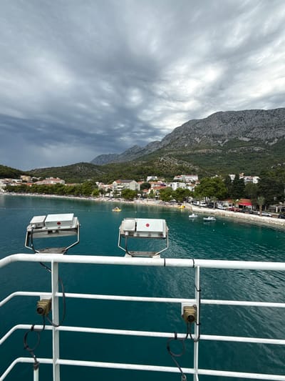 A photograph taken from the deck of a vessel, looking towards the coast of Drvenik, Croatia. The water in the foreground is a vibrant turquoise, with white railings and two large, metal floodlights visible. The floodlights have black cables attached and appear to be mounted on a metal frame. Beyond the railing and lights, a beach is visible, lined with trees and buildings. The beach is populated with numerous people, some of whom are sunbathing or swimming. Several small boats are anchored in the water. The town is nestled at the base of a steep, rocky mountain covered in dense green vegetation. The sky above is overcast with dramatic, dark clouds. The overall impression is of a coastal town on a cloudy day, viewed from the sea.