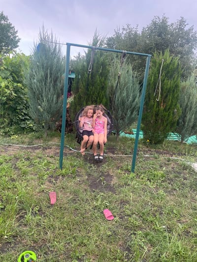 In this horizontal, full-shot image, two young girls are seated on a circular swing. The girl in front is wearing a pink swimsuit with a floral pattern and black cycling shorts. She has blonde hair and is looking down at her lap with her eyes closed, her mouth slightly open in what appears to be a contented or tired expression. Behind her, another girl in a pink tank top and black shorts is leaning back, resting her head on the front girl's shoulder. She has blonde hair and is looking towards the camera with a relaxed expression. Both girls are barefoot.
The swing is suspended from a green metal frame, which is visible in the center and left of the frame. The frame is sturdy and appears to be set up in a backyard or garden. The area surrounding the swing is covered in green grass and some patches of bare earth. Behind the swing and to the left, there are tall, dense evergreen trees, likely junipers, that create a lush, natural backdrop. To the right of the swing, the grass continues, with some scattered weeds and a small yellow flower.
In the background, behind the trees, a green corrugated metal fence is partially visible. Scattered on the grass to the right of the swing are two pink flip-flops and a small green object that could be a garden sprinkler or part of a toy. The lighting suggests it is daytime, with an overcast sky, as indicated by the diffused light and the soft shadows. The overall impression is one of a peaceful, outdoor setting, likely a backyard in Raslovka 1-Ya, Russia, where the girls are enjoying a moment of rest on the swing after playing.
