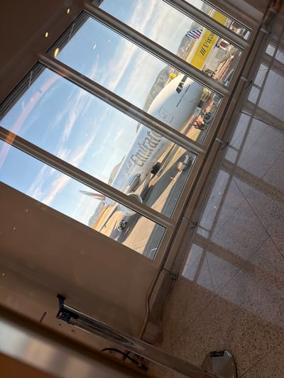 The image depicts an Emirates Boeing 777-300ER aircraft on the tarmac, viewed through the windows of what appears to be an airport terminal. The aircraft is being serviced by ground crew and equipment. A "Piraeus" sign is visible. The location is likely at Athens International Airport "Eleftherios Venizelos" in Spata-Loutsa Municipal Unit, Greece, given the presence of an Emirates aircraft and the visible "Piraeus" sign. No people are clearly visible to identify their actions and interactions. The environment appears to be a sunny day.