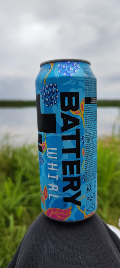 A close-up, eye-level shot captures a blue can of Battery energy drink, specifically the "Raspberry Whirl" flavor, resting on dark gray fabric, possibly clothing. The can's design features images of raspberries and abstract floral patterns in pink and yellow. Text on the can is partially legible, mentioning "LIMITED 500 ML" and various ingredient and nutritional information in multiple languages, including what appears to be Estonian and possibly other Baltic languages. The background is softly blurred, showing a calm body of water with a green grassy shoreline, indicative of an outdoor setting. The sky above is overcast and bright. The overall impression is that of a casual outdoor moment in a tranquil, natural environment, likely in the vicinity of Vītoliņi, Latvia.