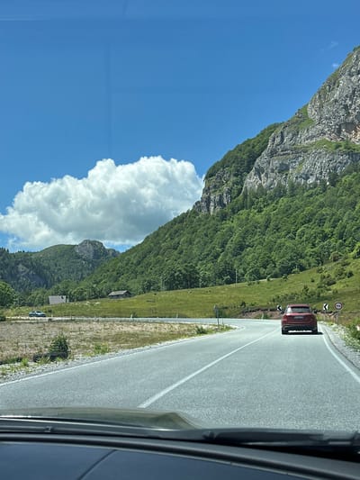 A red SUV is driving on a paved road through a mountainous landscape in Lubnice, Montenegro. The road curves to the right as it ascends. Steep, rocky mountains covered in dense green trees rise on the right side of the road. A large, fluffy white cloud is visible in the bright blue sky above the mountains. In the distance, on the left side of the road, there are rolling green hills with two small buildings, one with a dark roof and the other a lighter colored structure. Another car, a silver sedan, is parked on the left side of the road in the distance. The image is taken from inside another vehicle, with the dashboard and windshield wipers visible in the foreground.