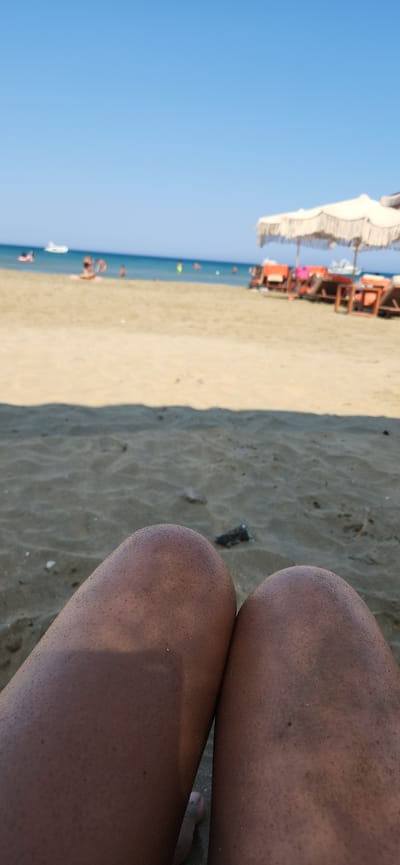 The image shows a close-up view of two legs and knees on a sandy beach, with the ocean and a beach scene in the blurred background. The legs are tanned and covered in fine sand. In the background, there are beach umbrellas, lounge chairs, and people enjoying the water. The sky is clear and blue. The location is specified as the city of Akrotiri, Cyprus.