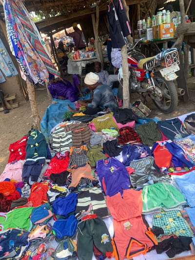 This is a medium-shot, high-angle photograph taken outdoors, likely in Boi A, Nigeria. The scene depicts a bustling open-air market stall overflowing with a colorful assortment of clothing. In the foreground, a large pile of folded fabrics, featuring vibrant geometric patterns and floral motifs, is displayed prominently.

Dominating the mid-ground is a motorcycle, its rear wheel and part of its frame visible, positioned amongst stacks of clothing. To the left, shelves are laden with numerous bottles, some appearing to be pesticides or agricultural products, with prominent text including "1L" and other illegible labels.

Several individuals are present in the scene. In the foreground, a person wearing a striped shirt is partially visible. In the mid-ground, a person in a purple garment is seen. Further back, a person in a white cap and another in a dark shirt are interacting near the shelves of bottles. The overall impression is that of a busy marketplace, possibly in the morning or afternoon given the natural lighting. The ground appears to be dirt. The arrangement of goods suggests a commercial activity of selling apparel and other wares.