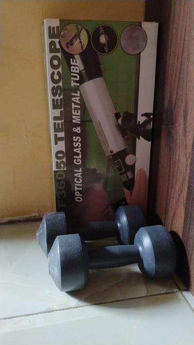 In a corner of a room with pale yellow walls and a light-colored tiled floor, a cardboard box for a "F36050 TELESCOPE OPTICAL GLASS & METAL TUBE" leans against a wooden surface. The box features images of celestial bodies and a bird. In front of the box, two dark gray dumbbells rest on the floor, positioned parallel to each other. The lighting suggests it is daytime, with shadows cast on the floor and wall. The scene appears to be indoors, possibly in a residential setting within Banjarsari, Indonesia.