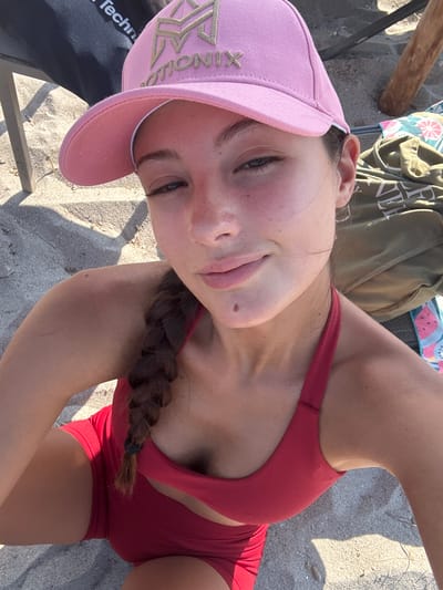 This is a close-up, upside-down selfie of a young person with light skin, taken outdoors on a sandy surface. The subject is wearing a red tank top or swimsuit top and red shorts. They are also wearing a pink baseball cap. A braid of dark brown hair is visible, extending over their shoulder. The background shows sand and a bit of a wooden object, possibly a piece of driftwood or furniture. There are some shadows suggesting it is daytime, likely sunny. On the pink cap, there is a logo with a star-like design and the word "MOTIONIX" in gold lettering. A black piece of clothing with white text "Techn" is visible at the bottom left edge of the frame. The scene appears to be a beach or a sandy area. The subject's expression is neutral.