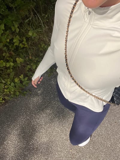 The image presents a downward-angled view of a person outdoors at night or in low light. The subject is wearing a white long-sleeved athletic top featuring a quarter-zip collar and thumbholes on the sleeves, combined with dark blue leggings. A gold-colored chain with black interwoven material, forming a strap, crosses the person's torso from the left shoulder (viewer's perspective) towards the right hip. A portion of a black quilted bag is visible on the right hip, attached to this chain. The person's left hand is positioned open by their side. The ground beneath is a dark, speckled asphalt or paved surface. To the left, dense green foliage, consisting of various plants and grasses, borders the path. The specific location within Skanderborg, Denmark, is not discernible from the image content.