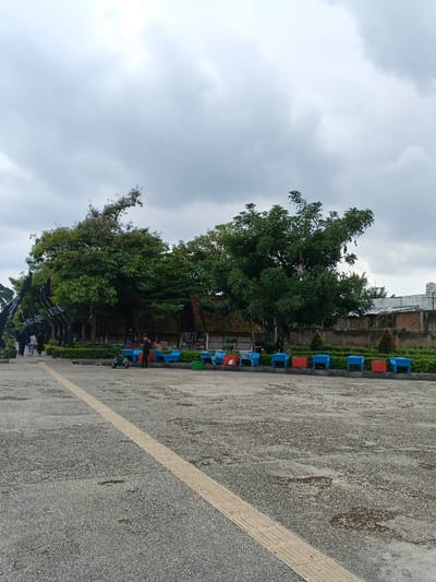 The image portrays an outdoor public area in thành phố Biên Hòa, Vietnam, under an overcast sky. The foreground features a broad, grey paved surface composed of small aggregate, bisected by a light-colored tactile paving strip running diagonally from the lower center to the mid-right.
In the midground, a dense line of green trees extends across the frame. To the far left, partially obscured by trees, a dark, curved structure, possibly an archway or decorative element, is visible. Several indistinct figures are seen walking in this area towards the background. Central to the midground, a rustic-style building with a thatched roof and wooden beam supports is situated among the trees.
To the right of the central building, a series of small, brightly colored rectangular stands or tables, predominantly blue and red, are arranged in a line along a low green hedge. One individual, dressed in dark clothing, stands near a blue stand, facing towards the rustic building. A small green wheeled cart is positioned on the pavement near this person. Additional figures are present near the colored stands, their actions not clearly discernible. Beyond the foliage and stands, a low brick wall is visible, with a larger, light-colored building in the far background. The sky is uniformly grey and heavily clouded, indicating diffused natural light.