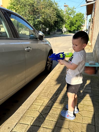 This image depicts an outdoor scene in Hadzhidimitrovo, Bulgaria, under clear, sunny conditions. A young male child, estimated to be between three and five years old, stands on a light-colored paver sidewalk, facing left. He has a dark, faded haircut with a distinct line shaved into the side. The child is wearing a white short-sleeved t-shirt, dark shorts, white socks, and blue Crocs. He is holding a blue and neon green water gun with both hands, aimed towards a light grey/silver sedan or hatchback partially visible on the left. Water streaks are evident on the car's front passenger window, indicating recent spraying, and the car's body appears dusty.
To the child's right, a textured concrete or stucco building wall is present, featuring a wooden overhang and a metal downspout. A small, light blue-topped concrete or stone stool with a terracotta pot beneath it rests against this wall. The paved sidewalk displays elongated shadows, suggesting a low sun angle from the right. In the background, beyond the car, a paved street is visible, bordered by dense green trees. A large stork nest is conspicuously positioned atop a utility pole above the trees. The sky is clear blue.