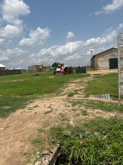 In Lau, Nigeria, on a bright, cloudy day, a scene unfolds with a focus on agricultural machinery and structures. A large, red and white combine harvester is positioned on the left side of the frame, facing right. To its right, a dark-colored agricultural trailer with a metal frame sits behind the harvester. Further to the right, a single-story building with a grey concrete exterior and green double doors dominates the background. The building appears to be a storage facility or workshop. The ground in the foreground is a mix of dirt and sparse green grass, with some debris scattered around. Beyond the machinery and building, there are several residential structures with pitched roofs, some featuring light blue roofing. A grey brick wall forms a boundary in the middle ground. A tall pole with a solar panel is visible near the building. A metal signpost with partial text is on the far right edge of the image. The overall environment suggests a rural or semi-rural setting within the city.