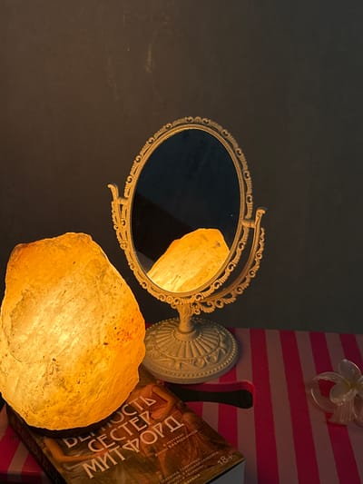 This media file captures a still life composition featuring several objects illuminated by a warm, artificial light source, likely a salt lamp which is prominently displayed in the foreground. The salt lamp is an irregularly shaped, translucent, yellowish-orange rock emitting a soft glow, casting its light on a vintage-style ornate mirror with a cream-colored frame. The mirror reflects a portion of the salt lamp.
In the background, a book with a title in Cyrillic script is partially visible. The visible text on the book includes "18+", "ДЕЛО", "СЕСТЕР", "МИТФОРД", and mentions names like "АННЫ БАБЯШКИНОЙ" and "AMERICA". This suggests the book is a translated work, possibly a novel.
The setting appears to be indoors, with a dark, textured wall on the left and a surface with pink and white stripes on the right, possibly a decorative backdrop or furniture. A small, translucent white flower ornament is affixed to the striped surface. The overall mood conveyed is one of quiet intimacy, with the warm light creating a cozy and perhaps contemplative atmosphere. There are no people or animals visible. The time of day and weather are not discernible from the image. The location context provided is Tliustenkhabl', Russia, which might be the context in which this image was taken.