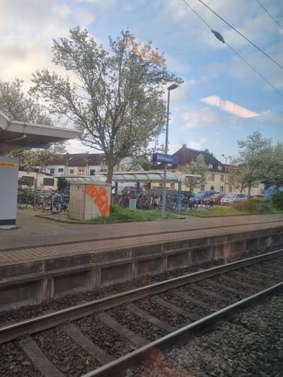 The image shows the train station "Bühl (Baden)" in Bühl, Germany, viewed from a moving train. The station platform is visible on the right, with railway tracks next to it. The station building is partially obscured by a tree, but the station sign "Bühl (Baden)" is visible. Several bicycles are parked in bike racks near the station. A DHL parcel station and an electrical box with graffiti are also visible. The sky is partly cloudy.