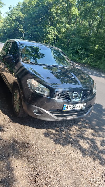 The image depicts a black Nissan Qashqai parked on the side of a paved road, with its right side facing the viewer. The car is positioned on a gravel and dirt shoulder adjacent to a dense forest. The road appears to be in a rural or semi-rural setting, likely within the city of Ivanhorod, Ukraine, given the Ukrainian license plate. The license plate reads "AX 5823 IM". The car's headlights and grille are visible, and the sun is casting shadows on the ground through the trees. The environment is characterized by lush green foliage and clear, sunny weather. No people are visible in the frame, and the car appears to be stationary. The overall scene is one of a parked vehicle in a natural, wooded environment.