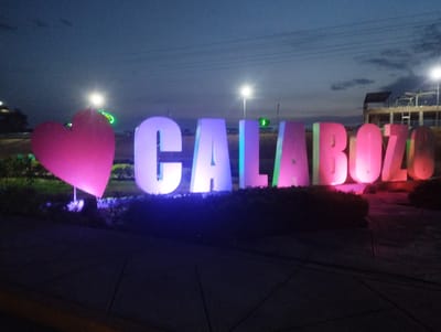 The image shows a brightly lit sign that spells out "CALABOZO" with a heart symbol replacing the first letter "C", located in Villa de Todos los Santos de Calabozo, Venezuela. Each letter is individually illuminated, showcasing different colors including pink, purple, and blue. The sign is set within a garden area with low bushes and greenery. In the background, streetlights are visible against a twilight sky, and a building structure can be discerned. The foreground is a paved surface.