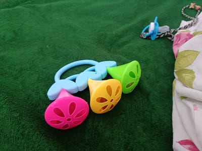 The image displays a collection of baby toys and a pacifier lying on a dark green, plush surface, likely a blanket or carpet. In the foreground, a set of three colorful plastic rattles are arranged. The leftmost rattle is bright pink and shaped like a bell with a flower-like pattern cut into its surface. To its right is a yellow rattle, also bell-shaped with a similar floral cutout pattern. The third rattle, to the right of the yellow one, is bright green and also features a perforated, bell-like design. These rattles are connected to a light blue, flexible plastic ring with leaf-shaped attachments. In the upper right quadrant of the image, a blue and white pacifier is visible, attached to a patterned black and white cord with small wooden beads. A white fabric with a floral pattern, featuring pink and green leaves, is partially visible in the far right of the frame. The overall scene is indoors, with the textured green surface suggesting a soft, possibly domestic environment. There is no indication of the city of Arkadag, Turkmenistan in the image itself.