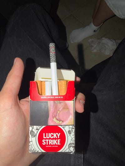 A hand holds a pack of Lucky Strike cigarettes. A single cigarette is pulled slightly out from the pack. The pack has a dental warning image on it. The scene is likely indoors, given the lighting and the presence of clothing on what appear to be limbs. There is a white sock on the left and some white material (possibly a crumpled garment) nearby. The image provides insufficient data to determine the precise location in Punta Umbría, Spain.