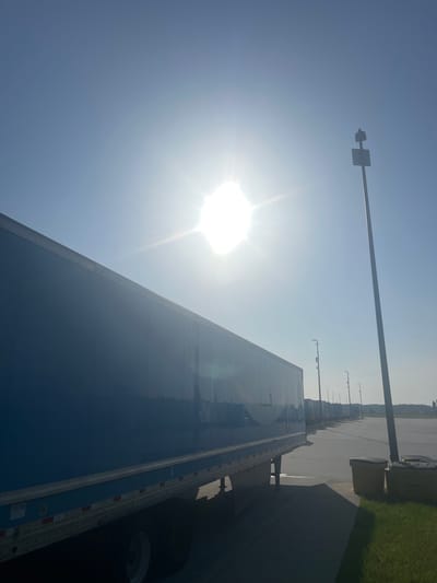 The image shows a bright, sunny day with the sun high in the sky, causing lens flare. In the foreground, the side of a blue semi-trailer is visible, angled towards the right of the frame. The trailer is parked in what appears to be a large lot, possibly a truck stop or distribution center. Several other semi-trailers are parked in a line behind the first one, receding into the distance. A tall pole with a sign and what looks like a siren or speaker is visible on the right side of the image. Further to the right, two large yellow trash bins are situated on the pavement next to a grassy area. The overall environment suggests an industrial or logistical setting. This scene is likely located in Pooler, Georgia, United States, which is known for its significant trucking and logistics presence.