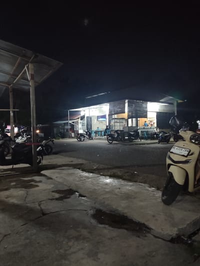 The image depicts an outdoor scene at night, likely a roadside eatery or shop in Cotglumpang, Indonesia. The setting is illuminated by artificial lights from the building and possibly streetlights.

In the foreground, several motorcycles are parked. One cream-colored scooter is prominently featured on the right side, with its license plate clearly visible: "BL 4326 ZBM" and "05-30". To the left, more motorcycles are parked under a corrugated metal roof supported by wooden and metal posts, creating a sheltered area.

The main building in the mid-ground appears to be a small establishment with large windows. Inside, several people are seated at tables, suggesting a dining or gathering activity. The interior is brightly lit, contrasting with the dark night sky. Some people are standing or moving around inside. The exterior of the building features a mix of open windows and covered sections, with blinds or screens visible.

The ground in the foreground is a paved surface, possibly concrete, showing signs of wear and tear, including cracks and damp patches that reflect the artificial light, indicating recent rain or a wet surface.

The overall mood is casual and functional, typical of a local establishment open in the evening. The time of day is clearly night. There are no obvious indicators of weather conditions beyond the wet ground. The presence of parked motorcycles and people gathered suggests a relatively active, albeit quiet, nighttime scene.