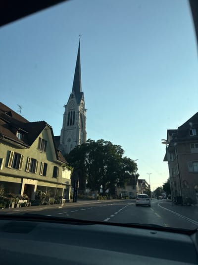 The image, taken from inside a vehicle, captures a view of a street in Amriswil, Switzerland. The scene is oriented vertically, suggesting the photo was taken while driving. The sky is clear and bright blue, indicating daytime with fair weather.

Dominating the left side of the frame is the tall, pointed spire of a church, its intricate architectural details clearly visible against the sky. Below and to the right of the church spire, a large, leafy green tree stands prominently. Several buildings are visible along the street, some with multi-story facades and windows. One building in the foreground has a pale green exterior with rows of windows and dark shutters. Another building further down the street appears older, with darker stonework.

A paved road runs through the scene, with white dashed lines marking the lanes. A white car is driving on the road, heading away from the viewer. The reflection or shadow of the vehicle from which the photo is taken is visible on the right side of the frame, a dark, textured surface.

There is a sign visible on one of the buildings, reading "SZENA NAILS". The overall impression is of a quiet, residential street in a European town on a sunny day. The time of day appears to be late morning or early afternoon, given the bright sunlight and the angle of the shadows, though this is not definitively clear due to the image orientation.
