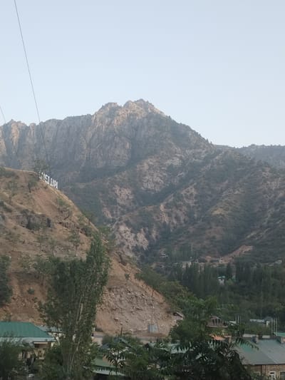This media file captures a scenic view of a mountainous landscape in Pughus, Tajikistan. The scene is dominated by rugged, rocky mountains that rise majestically against a pale, clear sky, suggesting a time of day around sunrise or sunset. The terrain is a mix of barren slopes and areas covered with sparse vegetation and trees.
On the left side of the frame, a large white sign is prominently displayed on a hillside. The text on the sign is in Cyrillic script and reads "САМЕДАРА". This appears to be a place name or an announcement related to the location. Below this sign, the hillside transitions into a more vegetated area with a few scattered trees.
Further down the slope and in the valley below, glimpses of a village or settlement can be seen. Several houses with green roofs are visible, nestled amongst the trees. The presence of these buildings and the cultivated appearance of some areas indicate human habitation and activity in the region.
The overall atmosphere is serene and peaceful, with the natural grandeur of the mountains being the primary focus. The weather appears clear, with no indication of rain or clouds. The lighting suggests it is either early morning or late afternoon, casting soft light on the landscape and creating subtle shadows. The image evokes a sense of natural beauty and rural tranquility.
