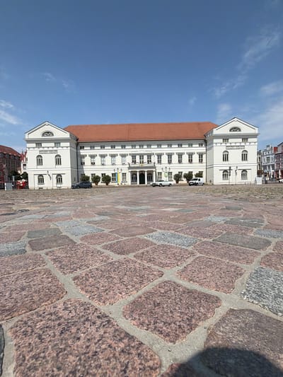This is a wide-angle, low-angle exterior shot of the historic Wismar City Hall, located in the Market Square of Wismar, Germany. The building is a large, white neoclassical structure with a prominent red-tiled roof and symmetrical pedimented facades. The foreground is dominated by a vast expanse of cobblestones, varying in color from pinkish-red granite to gray stone, laid in a somewhat irregular pattern. The sky is a clear, vibrant blue with a few wispy white clouds. In front of the building, a few cars are parked on the cobblestone square, including a dark gray SUV and a white van. There are also potted plants and flags visible near the building's entrance. The overall impression is one of a sunny day in a well-maintained historical city center.