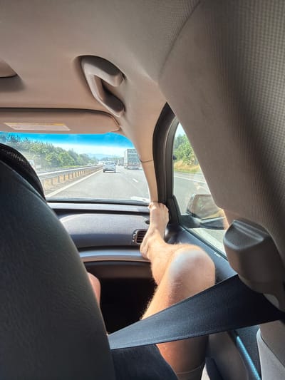 This media file captures an interior view from a vehicle traveling on a highway, as indicated by the road, guardrail, and passing cars visible through the window. The scene is shot from the perspective of someone in the passenger seat, looking forward and slightly upwards. A person's bare leg and foot are prominently displayed on the dashboard, suggesting a relaxed or informal posture during the journey. The sun is shining, creating bright conditions and casting some shadows, hinting at daytime. The sky is a clear blue. The interior of the car features fabric upholstery on the ceiling and a black seatbelt. A grab handle is visible on the ceiling. The overall impression is one of a road trip on a sunny day. There is no visible text in the image.
